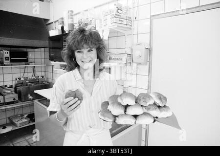 Marchel Flowers Providing Sandwiches Bolletje Eatery IJmuiden Brood IJmuiden the Netherlands, Whizgle News, Dutch Desk, Niederlande, 1950 - 2000 am 15-08-1988. Dies sind die Elemente im Bild. Die Szene zeigt eine fröhliche Person, die in einer hellen und gut beleuchteten Küchenumgebung steht. Sie hat voluminöses, welliges Haar, das ihr Gesicht umrahmt und ein freundliches und einladendes Verhalten ausstrahlt. In der einen Hand hält sie eine einzelne Rolle, während ihr anderer Arm nach außen reicht und eine Platte präsentiert, die mit mehreren ähnlich gebackenen Waren gefüllt ist – goldbraune Brötchen, die ein warmes und appetitliches Aussehen verleihen. Die Stockfoto