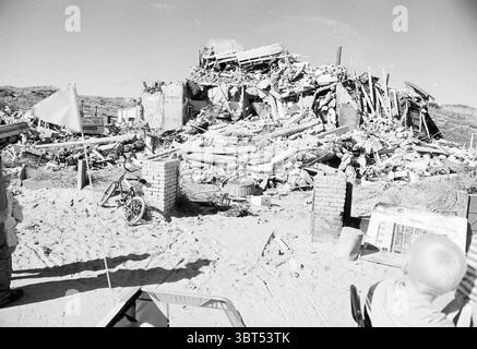 Abriss von Emmas Haus, Whizgle News, Dutch Desk, Niederlande, 1950 - 2000 am 31-08-1993. Die Abbildung zeigt diese Themen. Die Szene zeigt die Folgen der Zerstörung, wo eine große, zusammengebrochene Struktur den Hintergrund dominiert. Die Überreste von Wänden, Balken und Trümmern bilden ein chaotisches Mosaik aus Materialien, die über den Sandboden verstreut sind. Staub und Schutt fügen eine Schicht der Verwüstung hinzu. Im Vordergrund sind einige Personen zu sehen, darunter ein Kind mit einem hellen Hemd, das aufmerksam auf die Stelle blickt. Auf der einen Seite neigt sich ein kleines, verwittertes Fahrrad gegen eine Ziegelstütze, sein verblichenes c Stockfoto