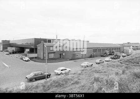 Unternehmen/Lager, Whizgle News, Dutch Desk, Niederlande, 1950 - 2000 im Jahr 1977. Das Bild enthält diese Themen. Die Szene zeigt ein Industriegebiet, das durch eine Mischung von Gebäuden und Fahrzeugen gekennzeichnet ist. Im Vordergrund befindet sich eine schmale Straße, die von mehreren Autos gesäumt ist, von denen alle ältere Modelle zu sein scheinen, was auf eine Zeit vor einigen Jahrzehnten hindeutet. Die Fahrzeuge werden parallel zum Bordstein geparkt, wodurch ein Gefühl der Ordnung auf dem offenen Raum entsteht. Auf der linken Seite fällt ein zweistöckiges Backsteingebäude auf, das eine einfache, schlichte Fassade mit ein paar Fenstern aufweist, was darauf hinweist, dass es als Büro dienen kann Stockfoto