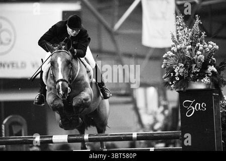 Jumping Lisse, Whizgle News, Dutch Desk, Niederlande, 1950 - 2000 am 22-01-1994. Dies sind die Themen im Bild. Ein Reiter navigiert geschickt mit einem Pferd über eine hochragende Schanze. Die Muskeln des Pferdes sind angespannt und zeigen seine Sportlichkeit, während es anmutig durch die Luft schwingt. Der Fahrer trägt formelle Reitkleidung, die sich durch eine taillierte Jacke und einen Helm auszeichnet, was ein Gefühl von Konzentration und Entschlossenheit widerspiegelt. Um die Szene herum sind eine Vielzahl von bunten Blumen kunstvoll in einem großen Display in der Nähe des Sprungsockels angeordnet, die einen lebhaften Kontrast zu den unterwürfigen bilden Stockfoto