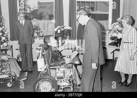 Installation von Bürgermeister Machielsen Zandvoort Mayors, Whizgle News, Dutch Desk, Niederlande, 1950 - 2000 am 05-10-1977. Das Bild enthält diese Themen. In der Szene interagiert eine Gruppe von vier Personen innerhalb eines gut beleuchteten Raumes, dessen Wände mit Dekorationen und Topfblumen geschmückt sind, die trotz der Grautöne ein Gefühl von Wärme vermitteln. Der Boden ist strukturiert, möglicherweise gekachelt oder gemustert, was zu einer strukturierten und dennoch einladenden Umgebung beiträgt. In der Mitte greift ein Mann im Rollstuhl mit einer anderen Person in Kontakt, die in der Nähe steht. Die sitzende Person wirkt entspannt, in dunkler Kleidung gekleidet Stockfoto