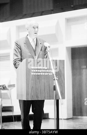 Man hält eine Rede vor dem Theater/Theatersaal auf dem Sprechtisch: stiftung für Berufsausbildung im Bauunternehmen, Whizgle News, Dutch Desk, Niederlande, 1950 - 2000 am 08-1971. Das Bild enthält diese Themen. Die Szene spielt in einem geräumigen Auditorium mit modernem, minimalistischem Design. Ein markantes Holzpodium steht in der Mitte, mit klaren Linien und einer glatten Oberfläche, die einen dezenten Glanz reflektiert. Hinter dem Podium besteht die Kulisse aus großen, geometrischen Formen in hellen Panels, die eine ausgewogene und strukturierte visuelle Umgebung schaffen. Die IND Stockfoto