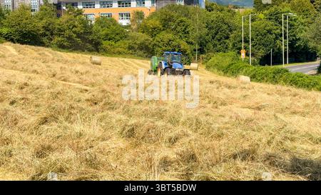 Pontypridd, Wales, Vereinigtes Königreich - 12. Juli 2025: Schlepper schleppen eine Ballenpresse, um große Rundballen aus getrocknetem Gras auf einem Feld herzustellen. Stockfoto