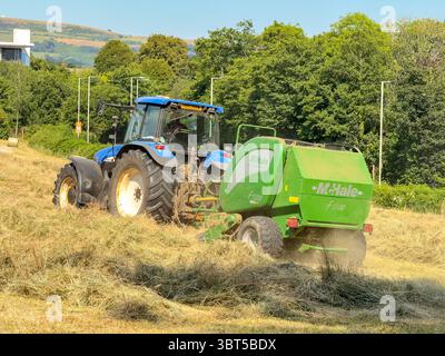 Pontypridd, Wales, Vereinigtes Königreich - 12. Juli 2025: Schlepper schleppen eine Ballenpresse, um große Rundballen aus getrocknetem Gras auf einem Feld herzustellen. Stockfoto