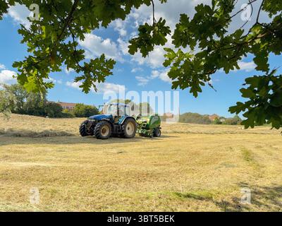 Pontypridd, Wales, Vereinigtes Königreich - 12. Juli 2025: Schlepper schleppen eine Ballenpresse, um große Rundballen aus getrocknetem Gras auf einem Feld herzustellen. Stockfoto