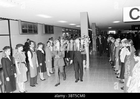 Eröffnung C-Pier Schiphol von Prinz Claus Schiphol 13-12-1987 Schiphol, Whizgle News, Dutch Desk, Niederlande, 1950 - 2000 am 13-12-1987. Das Bild enthält diese Themen. Die Szene fängt eine geschäftige Innenumgebung mit einem langen Korridor ein, der von Menschen auf beiden Seiten gesäumt ist. Die Architektur zeichnet sich durch schlanke, moderne Linien aus, die auf ein Design aus der Mitte des 20. Jahrhunderts hinweisen, mit einer minimalistischen Ästhetik. Auf der linken Seite steht eine Gruppe elegant gekleideter Individuen in ordentlicher Form. Sie tragen eine Vielzahl von maßgeschneiderten Outfits, überwiegend dunkle, formelle Kleidung, die einen Kontrast zu den helleren Tönen des Sur bildet Stockfoto