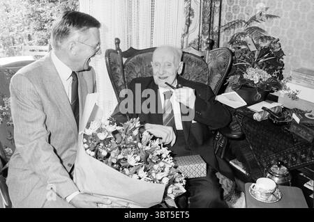 Grandpa Zaayer 106 Jahre alt Velsen-Noord Hundert Jahre Velsen-Noord, Whizgle News, Dutch Desk, Niederlande, 1950 - 2000 am 18-08-1983. Diese Themen sind in der Abbildung dargestellt. Die Szene zeigt zwei Männer in einem gemütlichen, warm dekorierten Zimmer. Ein Mann, in einem hellgrauen Anzug gekleidet, steht mit einem Blumenstrauß in den Armen, sein Gesicht leuchtet mit einem breiten Lächeln. Die Blumen sind eine lebendige Mischung aus Farben, die in hellen Rot-, Gelb- und Weißtönen gehalten sind und wunderschön in einer Papierverpackung angeordnet sind. Der ältere Mann sitzt bequem in einem großen, reich verzierten Sessel und hat einen gemütlichen Expressi Stockfoto