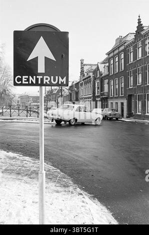 Einbahnschild Haarlem the Netherlands, Whizgle News, Dutch Desk, Niederlande, 1950 - 2000 im Jahr 1965. Diese Themen sind in der Abbildung dargestellt. In dieser Szene biegt sich eine Straße sanft nach links, wo ein weißer Oldtimer in Richtung Vordergrund steht. Das Fahrzeug wirkt klassisch, mit abgerundeten Kanten und einer markanten Form, die an das Design der Mitte des 20. Jahrhunderts erinnert. Um das Fahrzeug herum ist der Straßenbelag nass, wahrscheinlich aufgrund von neuem Regen oder schmelzendem Schnee. Das Licht reflektiert das Licht und verleiht der Oberfläche ein Gefühl von Glanz. Auf der rechten Seite sind die Gebäude eng aneinander gereiht, mit hohen, schmalen Fassaden wi Stockfoto