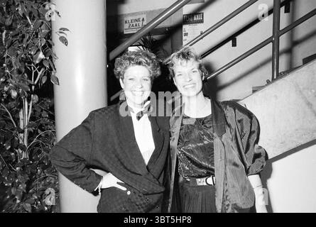Festiball Concertgebouw Football Haarlem, Whizgle News, Dutch Desk, Niederlande, 1950 - 2000 am 28-01-1989. Dies sind die Themen im Bild. In der Szene stehen zwei Frauen eng zusammen, die beide ein Gefühl von Selbstvertrauen und Kameradschaft ausstrahlen. Im Hintergrund befindet sich eine Treppe, die sich umgibt, was auf eine lebhafte Inneneinrichtung, möglicherweise auf einen Event- oder Versammlungsraum, hinweist. Eine Frau auf der linken Seite hat einen unverwechselbaren Haarstil, der durch voluminöse Locken gekennzeichnet ist, was eine verspielte und dennoch raffinierte Atmosphäre verleiht. Sie trägt ein maßgeschneidertes dunkles Outfit mit einer Fliege, die einen schicken, fast unauffälligen Look verleiht Stockfoto