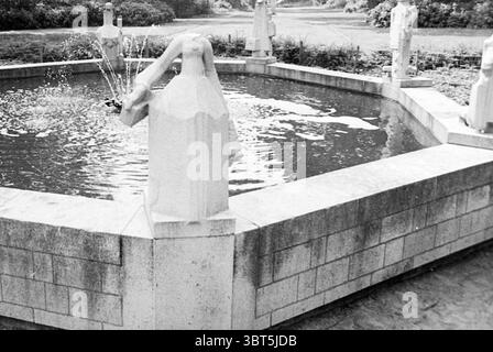 Statuen enthauptet am Rand eines Teiches Haarlem Oranjelaan, Whizgle News, Dutch Desk, Niederlande, 1950 - 2000 am 21.06.1973. Dies sind die Themen im Bild. Die Szene zeigt einen ruhigen Brunnen in einem gepflegten Garten. Der Brunnen selbst verfügt über ein kreisförmiges Becken aus Stein mit einer glatten, reflektierenden Oberfläche, die das umgebende Grün und den Himmel einfängt. Skulpturen zieren die Seiten des Waschbeckens, jede einzelne im Design, und verleihen der Einrichtung ein Gefühl von Kunstfertigkeit und Eleganz. Die Charaktere sind zwar nicht identifizierbar, sind aber so angeordnet, dass sie auf Mo hinweisen Stockfoto