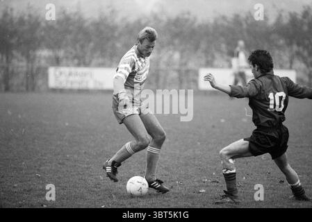 Football Velsenoord - Kennemeren Velsen, Whizgle News, Dutch Desk, Niederlande, 1950 - 2000 am 09-11-1990. Das Bild enthält diese Themen. In einer dynamischen Outdoor-Umgebung spielen zwei Spieler einen intensiven Moment auf einem grasbewachsenen Feld, in dem sich die Atmosphäre mit Wettbewerbsenergie aufgeladen fühlt. Die Szene ist von einem subtilen Nebel umhüllt und verleiht der Umgebung eine leicht ätherische Qualität. Der erste Spieler, der in einem hellen, mit Streifen verzierten Trikot gekleidet ist, ist fokussiert und agil, mit einem Fuß, der über einem weißen Fußball schwebt. Seine Shorts entsprechen der Farbe seines Trikots und schaffen so ein C Stockfoto