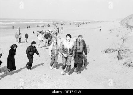 Winter Walk Kurier Haarlems Dagblad etc. Und IJmuider Courant und Koeri Walking, Whizgle News, Dutch Desk, Niederlande, 1950 - 2000 am 25-02-1990. Diese Themen werden in der Abbildung angezeigt. Die Szene entfaltet sich an einem Sandstrand, wo sich eine vielfältige Menschenmenge versammelt, die sich auf die ruhigen Wellen des Ozeans zubewegt. Die Küste erstreckt sich bis in die Ferne und trifft auf einen sanften Horizont, an dem der Himmel leicht auf das Wasser trifft. Die Leute tragen eine Mischung aus lässiger Kleidung, die für einen kühlen Tag am Meer geeignet ist, mit einigen sportlichen Windbreakern und anderen in bequemeren Lagen. Die Farbpalette ist Do Stockfoto