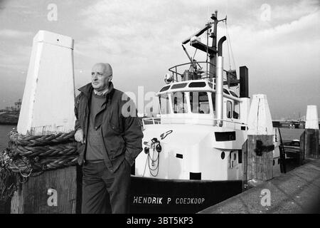 Mr. Boudesteijn 40 Jahre alt. Abschleppdienst, Whizgle News, Dutch Desk, Niederlande, 1950 - 2000 am 19-10-1994. Das Bild enthält diese Themen. Im Hafen steht ein Mann selbstbewusst neben einem stabilen, vertäuten Schiff. Er trägt praktische Kleidung, die auf eine Affinität zur maritimen Umwelt hindeutet, mit einer entspannten Haltung, die eine Vertrautheit mit der Umgebung impliziert. Die Farben in der Szene sind gedämpft, dominiert von Grau- und Schwarztönen, die charakteristisch für eine monochrome Ästhetik sind, die die ernste und kontemplative Stimmung verstärkt. Der Hintergrund verfügt über ein robustes Dock Stockfoto