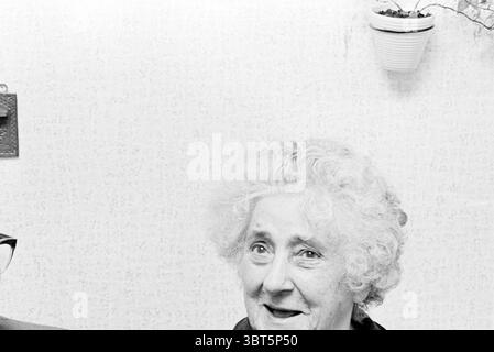 Porträt älteres Paar., Whizgle News, Dutch Desk, Niederlande, 1950 - 2000. Das Bild enthält diese Themen. In einem sanft beleuchteten Innenraum strahlt eine Frau mit flauschigen, weißen Locken Wärme und Zugänglichkeit aus. Ihr Gesicht ist mit sanften Falten geschmückt, die eine Geschichte eines gelebten Lebens erzählen, und ihr Ausdruck strahlt Freude aus, geprägt von einem breiten Lächeln. Der Hintergrund besteht aus einer strukturierten Wand, die hell ist und einen subtilen Kontrast zum Motiv schafft. Topfpflanzen, die möglicherweise in weißen Behältern platziert werden, hängen von der Decke ab und verleihen der Szene einen Hauch von Natur. Ihr grünes Lev Stockfoto