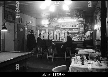 Family de Boer im Café Brabant Café, Whizgle News, Dutch Desk, Niederlande, 1950 - 2000 am 25-12-1960. Das Bild enthält diese Themen. Die Szene zeigt eine gemütliche, schwach beleuchtete Bar mit nostalgischer Atmosphäre. Im Vordergrund befindet sich ein großer Billardtisch auf einer Seite, dessen Grün sich leicht abgenutzt anfühlte, was darauf hindeutet, dass viele Spiele gespielt wurden. Die Wände sind mit verschiedenen Dekorationen verziert, die dem Raum Charakter und Wärme verleihen. An der Bar sitzt eine Gruppe von fünf Personen auf hohen Holzhockern, um sich zu unterhalten. Sie sind in Kleidung aus der Mitte des 20. Jahrhunderts gekleidet, was auf eine formelle oder halbförmige Form hindeutet Stockfoto