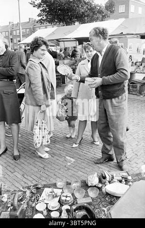 Market Vendor Market, Whizgle News, Dutch Desk, Niederlande, 1950 - 2000 am 10-09-1964. Diese Themen sind in der Abbildung dargestellt. Die Szene zeigt einen belebten Markt mit einer Vielzahl von Gegenständen, die auf Tischen und dem Boden liegen. Im Vordergrund präsentiert ein Mann einer Frau, die in der Nähe steht, animiert ein Objekt. Sie trägt einen hellen Mantel und trägt eine gemusterte Handtasche, während ein kleines Kind, das ihren Rock umklammert, neugierig auf die ausgestellten Gegenstände blickt. Das Kind trägt ein schlichtes Outfit, das sich in die zwanglose, aber lebendige Atmosphäre einfügt. Um diese Interakti herum Stockfoto