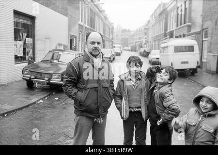 Herr Sevil Gemeindeentwickler Frans Halsbuurt Ausländer Nachbarschaften Gemeindezentren örtliche Geschäfte Trainingszentren, Whizgle News, Dutch Desk, Niederlande, 1950 - 2000 am 24-11-1983. Dies sind die Elemente im Bild. Die Szene zeigt eine Straßenansicht, die einen Moment in der Zeit festhält. Im Vordergrund stehen vier Individuen, drei Kinder und ein Erwachsener, der sich leicht von der Gruppe abhebt, alle in einer lässigen Anordnung. Die Kinder zeigen fröhliche Ausdrücke, wobei einer spielerisch eine Geste zur Kamera macht, während die anderen herzlich lächeln. Der Mann, Dres Stockfoto