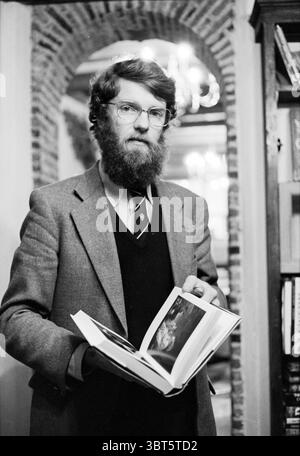 Porträt Heer de Vries Junior (Buchhandlung de Vries) Bücher und Buchhändler Porträt Haarlem the Netherlands, Whizgle News, Dutch Desk, Niederlande, 1950 - 2000 am 13-03-1980. Diese Themen werden in der Abbildung angezeigt. In einem warm beleuchteten Innenraum steht ein Mann prominent und hält ein offenes Buch in der Hand. Er hat einen Vollbart und trägt eine runde Brille, die seinen nachdenklichen Ausdruck ergänzt. Seine Kleidung besteht aus einem grauen Blazer über einem dunkleren Hemd, gepaart mit einer Krawatte, was auf ein Gefühl professioneller Formalität hindeutet. Der Hintergrund bietet eine gemütliche Atmosphäre mit Ziegelwänden, die Textur und Krieg verleihen Stockfoto