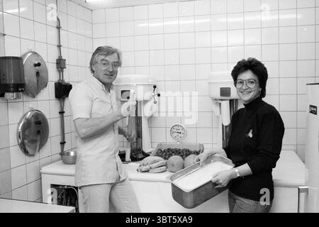 Eisdiele Tremonti Grote Houtstraat Eislaufbahn Eishockey Haarlem Grote Houtstraat Niederlande, Whizgle News, Dutch Desk, Niederlande, 1950 - 2000 am 23-06-1988. Das Bild enthält diese Themen. In einer hellen, gekachelten Küche arbeiten zwei Personen mit der Zubereitung von Speisen. Die Szene zeichnet sich durch einen klaren, weißen Hintergrund aus, der von großen, quadratischen Kacheln dominiert wird, die die obenliegende fluoreszierende Beleuchtung reflektieren und zu einer lebendigen und doch klinischen Atmosphäre beitragen. Links steht ein Mann lächelnd und hält einen Becher in einer Hand. Er hat kurze, ordentlich gestylte Haare und Stockfoto