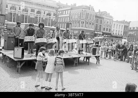 Music on the Grote Markt Music Haarlem Grote Markt the Netherlands, Whizgle News, Dutch Desk, Niederlande, 1950 - 2000 am 02-08-1974. Das Bild enthält diese Themen. Die Szene spielt auf einem lebhaften Platz im Freien, der voller Aktivität ist. Eine große hölzerne Plattform dominiert den Vordergrund, wo eine lebhafte Aufführung stattfindet. Auf der Bühne spielt eine Band von Musikern glänzende Stahltrommeln, die ihre rhythmischen Talente unter Beweis stellen. Die Musiker sind in einer Mischung aus lässiger und farbenfroher Kleidung gekleidet, die ein Gefühl von Spaß und Energie ausstrahlt. Vor der Bühne steht eine kleine Gruppe von Kindern verzaubert. Das sind sie Stockfoto