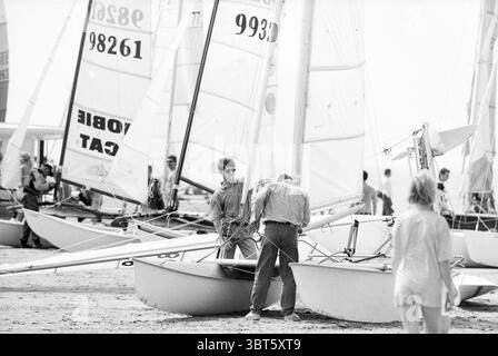 Katamarane Z'voort Zandvoort, Whizgle News, Dutch Desk, Niederlande, 1950 - 2000 am 13-05-1994. Das Bild enthält diese Themen. Die Szene fängt eine lebhafte Strandkulisse voller Aktivität ein. Im Vordergrund sind zwei Personen an der Vorbereitung eines kleinen Segelschiffs beteiligt. Eine Person mit stilvoller Sonnenbrille und entspanntem Auftreten kümmert sich um die Takelage, während die andere in einem leichten Hemd die Ausrüstung des Katamarans anpasst. Um sie herum liegen mehrere Boote im Sand, deren schlanke Rümpfe einen Hauch des hellen Tageslichts reflektieren. Die Segel sind PR Stockfoto