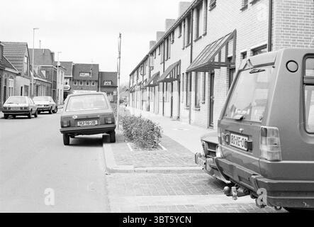 Autos teilweise auf dem Bürgersteig geparkt, Whizgle News, Dutch Desk, Niederlande, 1950 - 2000 am 27-05-1983. Diese Themen werden in der Abbildung angezeigt. Die Szene zeigt eine ruhige Straße, die von bescheidenen Wohnhäusern gesäumt ist, die alle mit einer einheitlichen Ziegelfassade gebaut sind. Die Gebäude sind zwei Stockwerke hoch und verfügen über symmetrische Fenster und schräge Dächer, die der Umgebung einen Hauch von Harmonie verleihen. Die Architektur deutet auf eine geplante Gemeinde hin, in der die Gebäude ähnliche Designelemente wie Holzläden und Vorveranden teilen. Im Vordergrund parken zwei Autos entlang der Bordsteinkante. Einer ist ein Kompaktmotor Stockfoto
