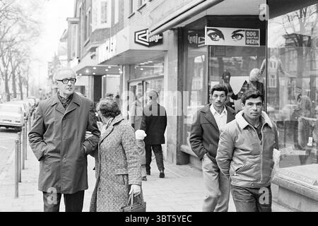 Käufer in einer Einkaufsstraße im Hintergrund eine Vroom & Dreesman-Filiale, Whizgle News, Dutch Desk, Niederlande, 1950 - 2000 am 02-1971. Dies sind die Themen im Bild. Die Szene fängt eine geschäftige Straße ein, in der eine Vielzahl von Menschen an ihren täglichen Routinen beteiligt ist. Auf der linken Seite steht ein älterer Herr mit seiner Brille und einem langen, maßgeschneiderten Mantel, der einen Hauch von Raffinesse ausstrahlt. Er geht an der Seite einer Frau mittleren Alters in einem strukturierten, dunklen Mantel, deren Haltung auf ein Gespräch oder einen gemeinsamen Moment hinweist. Im Hintergrund schlendern mehrere Männer beiläufig. Einer ist dr. Stockfoto