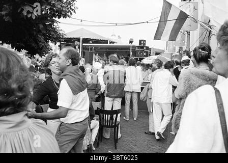 Jazz Zandvoort + Organisation Jazz Zandvoort Jazz Zandvoort Zandvoort Zandvoort, Whizgle News, Dutch Desk, Niederlande, 1950 - 2000 am 04-08-1979. Die Abbildung zeigt diese Themen. Die Szene fängt ein lebhaftes Treffen im Freien mit einer vielfältigen Menschenmenge ein, die Gespräche führt und ihre Umgebung genießt. Im Vordergrund steht ein Paar nahe beieinander, tanzt oder schwingt zur Musik, wobei seine Ausdrücke Freude und Verbundenheit zeigen. Die Frau hat lockiges Haar und trägt eine Brille, während der Mann ein helles Hemd und eine Hose trägt. Die Umgebung ist geschäftig und es kommen zahlreiche Personen zusammen Stockfoto