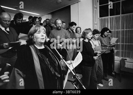 Singen Sie Protest von Nachbarschaftsräten im Rathaus mit Bürgermeister Schmitz Haarlem the Netherlands, Whizgle News, Dutch Desk, Niederlande, 1950 - 2000 am 24.11.1991. Das Bild enthält diese Themen. In einem geräumigen Innenbereich versammeln sich verschiedene Gruppen von Einzelpersonen, die eine chorartige Versammlung bilden. Im Vordergrund steht eine Frau, die selbstbewusst an einem Mikrofon steht. Ihr Ausdruck ist fokussiert und leidenschaftlich. Sie trägt dunkle Gewänder und verleiht der Szene einen Hauch von Zeremonie. Um sie herum stehen mehrere Personen in einem Halbkreis, jeder hält Blätter mit Papier, vermutlich Texte oder Stockfoto