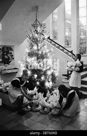 Home Schule Verpackung Weihnachtsmaterial Weihnachten Weihnachtsdekorationen Weihnachtsdekorationen Weihnachtsdekorationen Schulen, Whizgle Nachrichten, Dutch Desk, Niederlande, 1950 - 2000 auf 20-12-1962. Dies sind die Elemente im Bild. Die Szene spielt drinnen und zeigt einen großen, wunderschön geschmückten Weihnachtsbaum, der in der Ecke steht. Der Baum ist mit Lichtern geschmückt, die seine Äste sanft beleuchten und eine warme und festliche Atmosphäre schaffen. Die Basis des Baumes umgibt zahlreiche Geschenke, die sorgfältig angeordnet sind, mit festlichem Papier und Bändern, die dem sc einen Hauch von weiß und Farbe verleihen Stockfoto