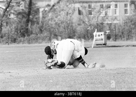 Bloemendaal-Rooswijk Baseball, Whizgle News, Dutch Desk, Niederlande, 1950 - 2000 am 11-05-1972. Das Bild enthält diese Themen. Die Szene fängt einen dynamischen Moment auf einem sonnendurchfluteten Baseballfeld ein. Zwei Spieler sind in Aktion, beide fokussieren sich intensiv auf den Ball, der nahe am Boden liegt. Ein Spieler, der in einer weißen Uniform mit schwarzen Akzenten gekleidet ist, liegt auf dem Boden und greift mit seinem Handschuh aus, um den Ball zu fangen. Seine Haltung ist dringend, sein Körper ist tief in die Erde und Schmutz wird um ihn herum geschleudert. Der zweite Spieler, der eine ähnliche Uniform trägt, ist in der Nähe. Stockfoto