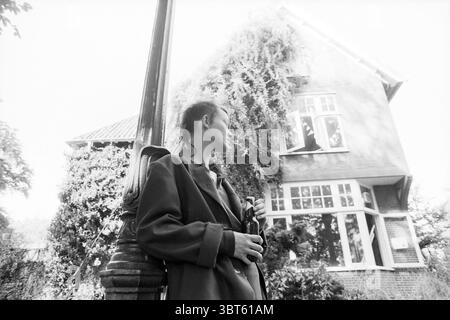 Kriemhilde Eviction, Whizgle News, Dutch Desk, Niederlande, 1950 - 2000 am 17-09-1991. Dies sind die Elemente im Bild. In einer urbanen Umgebung steht eine junge Frau neben einer alten Straßenlaterne, deren Silhouette vor dem Hintergrund eines mit Efeu bedeckten Hauses steht. Die Architektur des Hauses deutet auf einen klassischen Charme hin, mit großen Fenstern, die Licht- und Schattenflecken von den umgebenden Laubbäumen reflektieren. Die Frau trägt einen langen, übergroßen Mantel, der sich elegant um sie zieht, wobei der Stoff leicht von der Brise erfasst wird. Ihr Haar ist auf einzigartige Weise gestylt, vielleicht mit einem Pferdeschwanz Stockfoto