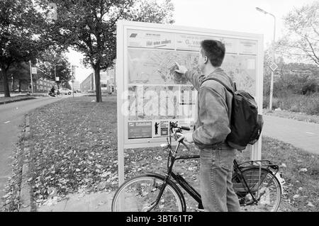 Karten der Gemeinde IJmuiden Velsen, Whizgle News, Dutch Desk, Niederlande, 1950 - 2000 am 07-10-1993. Das Bild enthält diese Themen. In einer monochromatischen Szene steht ein Mann neben einer großen Kartenanzeige, die in einer urbanen Umgebung deutlich positioniert ist. Er trägt lässig eine helle Jacke und eine dunklere Hose, die einen lässigen Stil verkörpert. Sein Rucksack liegt auf seinem Rücken, was auf ein Gefühl der Erkundung hindeutet. Der Mann zeigt mit einem fokussierten Ausdruck auf eine bestimmte Position auf der Karte und zeigt damit an, dass er beabsichtigt, durch das Gebiet zu navigieren. Die Karte selbst ist detailliert und zeigt verschiedene Elemente Stockfoto