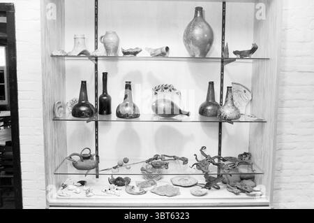 Alte Gegenstände in der Kutsche des Santpoort Zuid Provincial Hospital Antiquities, Whizgle News, Dutch Desk, Niederlande, 1950 - 2000 am 04-05-1983. Diese Themen werden in der Abbildung angezeigt. Die Szene zeigt eine Ausstellung verschiedener Artefakte, die auf einer Holzkonstruktion mit mehreren Regalen angeordnet sind. Die Objekte bestehen überwiegend aus Keramik und Glas und zeigen eine reiche Vielfalt an Formen und Größen. In den oberen Regalen stehen zahlreiche Flaschen und Vasen aufrecht, deren Oberflächen komplizierte Muster und Texturen zeigen, die auf historische Handwerkskunst hinweisen. Die Flaschen sind in unterschiedlichen Höhen erhältlich, einige davon haben sie Stockfoto