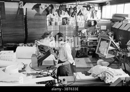 Siebdruckunternehmen Gejes v.d. Veen IJmuiden Printing House Printing IJmuiden the Netherlands, Whizgle News, Dutch Desk, Niederlande, 1950 - 2000 am 15-03-1982. Dies sind die Themen im Bild. Die Szene zeigt einen belebten Arbeitsbereich, der mit den Werkzeugen eines Siebdruckvorgangs gefüllt ist. Im Vordergrund konzentriert sich eine junge Frau intensiv auf ein Stück Stoff, während sie in einer großen Druckerei arbeitet. Ihr langes Haar fällt locker um ihre Schultern und sie trägt ein leichtes Hemd mit Knopfverschluss. Um sie herum ist der Raum lebendig mit einer Vielzahl von Farben und Formen. Verschiedene T-Shirts und Stoffe hängen ab Stockfoto