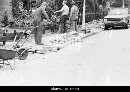 Beginn der Kabel-TV-Arbeit Heemskerk TV-Arbeit Heemskerk, Whizgle News, Dutch Desk, Niederlande, 1950 - 2000 am 11-08-1980. Die Abbildung zeigt diese Themen. Die Szene zeigt eine Straße, die im Bau ist. Im Vordergrund sind zwei Personen aktiv an der Arbeit beteiligt. Einer wird umgebeugt und bedient ein Werkzeug, während der andere in der Nähe steht und den Fortschritt beobachtet. Beide tragen praktische, strapazierfähige Kleidung, die für manuelle Arbeiten geeignet ist – Overalls und robuste Schuhe. Daneben befindet sich eine Schubkarre, die auf dem Pflastersteinpflaster positioniert ist, was auf eine laufende Aufgabe des Transportes von Material hindeutet. Das Rad Stockfoto