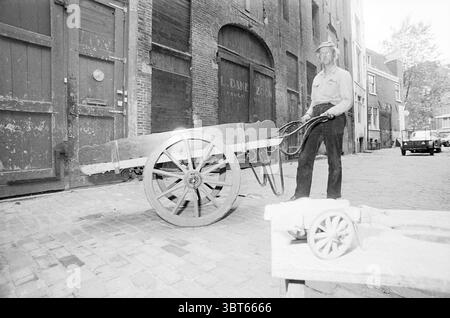 Guus de Mol mit Handkarren A'Dam Personen, Whizgle News, Dutch Desk, Niederlande, 1950 - 2000 am 19.07.1976. Dies sind die Elemente im Bild. In der Szene steht ein Mann neben einem Holzwagen, der große, robuste Räder und einen rustikalen Charme hat. Der Wagen befindet sich auf einer Kopfsteinpflasterstraße, was auf eine ältere Ära hindeutet. Der Mann trägt helle Kleidung und trägt einen Hut mit breiter Krempe, der einen Schatten auf sein Gesicht wirft. Seine Haltung ist entspannt und doch autoritativ, was darauf hindeutet, dass er mit der Umgebung vertraut ist. Die Kulisse zeigt verwitterte Backsteinhäuser, deren Texturen in der LIG sichtbar sind Stockfoto