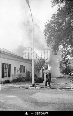 Fire Hartekamp Fire Brigade, Whizgle News, Dutch Desk, Niederlande, 1950 - 2000 am 01-09-1968. Das Bild enthält diese Themen. Die Szene fängt einen spannungsgeladenen, dramatischen Moment ein, der durch den rauschenden Rauch gekennzeichnet ist, der sich aus einem Gebäude links erhebt. Das Gebäude wirkt alt, mit seinen weiß getünchten Wänden und dunklen Fensterläden, von denen einige teilweise offen sind, wie in einem Alarmzustand. Der dichte und graue Rauch verdeckt einen Teil des Daches und deutet auf einen aktiven Brand hin, der sich im Inneren festgehalten hat. Im Vordergrund steht eine Figur ruhig auf der Straße. Im Dunkeln gekleidet, vielleicht schützend Stockfoto