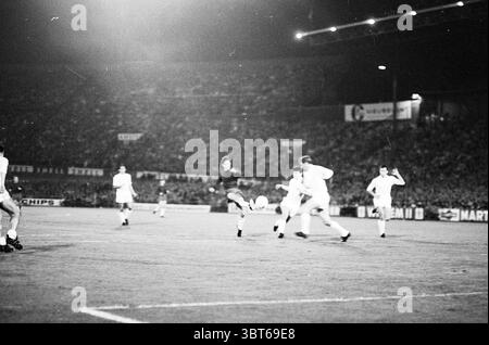Ajax - Real Madrid Football Ajax, Whizgle News, Dutch Desk, Niederlande, 1950 - 2000 am 20-09-1967. Die Abbildung zeigt diese Themen. Die Szene fängt einen dynamischen Moment während eines Fußballspiels ein. Im Vordergrund befindet sich ein Spieler in einer dunklen Uniform in der Mitte der Action und bereitet sich darauf vor, den Ball mit Präzision zu volleytieren. Der Ball ist hervorstehend, in der Luft schwebend und bildet einen Mittelpunkt der Bewegung. Um diese zentrale Figur herum sind zwei Spieler in weißen Trikots eng positioniert, die wahrscheinlich versuchen, sich gegen den Streik zu verteidigen. Ihre Ausdrücke spiegeln Konzentration und Entschlossenheit wider. Der Hintergrund ist gefüllt mit Stockfoto