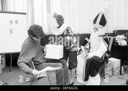 Sinterklaas kommt zu Besuch, Whizgle News, Dutch Desk, Niederlande, 1950 - 2000 am 12.04.1987. Das Bild enthält diese Themen. Die Szene fängt ein lebhaftes Ambiente mit festlicher Freude ein. Auf der einen Seite sitzt eine Person auf einem Holzstuhl, mit dem Rücken gewendet, in einen lässigen Strickpullover gekleidet. Ihre Haltung deutet auf eine Mischung aus Vorfreude und Schüchternheit hin, die sich leicht nach vorne neigt, während sie scheinbar etwas in ihren Händen untersuchen. Daneben spielt eine Frau in einem extravaganten Outfit mit bunten Mustern eine spielerische Interaktion. Sie trägt eine große Federboa Stockfoto