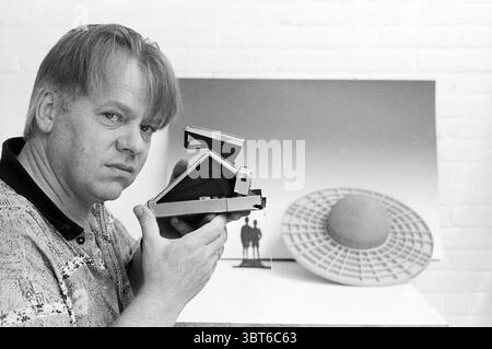 Piet van Leeuwen mit Polaroid Photography, Whizgle News, Dutch Desk, Niederlande, 1950 - 2000 am 19.07.1989. Diese Themen sind in der Abbildung dargestellt. In der Szene wird ein Mann deutlich im Vordergrund positioniert, der eine alte Kamera nahe an sein Gesicht hält. Er hat zerzauste Haare, die leicht über seine Stirn fallen, und sein Ausdruck ist ernst, aber besinnlich, was auf eine tiefe Auseinandersetzung mit seiner Aufgabe hindeutet. Sein Hemd hat ein auffälliges, farbenfrohes Muster, das einen Kontrast zu den gedämpften Tönen im Hintergrund bildet. Der Hintergrund zeigt eine große, flache Oberfläche, auf der ein gedrucktes Bild angezeigt wird. Diese ima Stockfoto