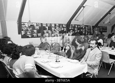 Essen mit Ausländern JHVU Foreign, Whizgle News, Dutch Desk, Niederlande, 1950 - 2000 am 07-03-1986. Die Abbildung zeigt diese Themen. Die Szene spielt in einem geräumigen Innenbereich mit einem großen, längs verlaufenden Tisch, umgeben von zahlreichen Personen. Die Atmosphäre wirkt warm, etwas nostalgisch, erfüllt von einem Gefühl der Gemeinschaft. Der Tisch ist mit einer weißen Tischdecke bedeckt und die Teller sind ordentlich angeordnet, was darauf hindeutet, dass eine Mahlzeit entweder serviert wird oder gerade beendet wurde. Gläser verschiedener Größen, die wahrscheinlich mit Getränken gefüllt sind, werden zeitweise entlang des Tisches platziert. Die Menschen, die sich hier befinden Stockfoto