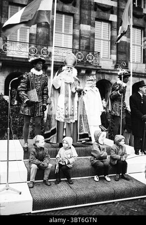Ankunft von Sinterklaas in Amsterdam Amsterdam Dam the Netherlands., Whizgle News, Dutch Desk, Niederlande, 1950 - 2000. Dies sind die Elemente im Bild. Auf einem belebten öffentlichen Platz sitzt eine Gruppe von Kindern auf einer gestuften Plattform, deren kleine Figuren in bunten Mänteln und Hüten verziert sind, was auf einen festlichen Anlass hinweist. Sie strahlen eine Mischung aus Neugier und Spannung aus, ihre Ausdrucksformen animiert, während sie die Figuren über ihnen beobachten. Im Vordergrund der Szene steht eine majestätische Figur in lebendigen, reichen Gewändern, verziert mit kunstvollen Mustern und einem hohen, spitzen Hut, der mit g glitzert Stockfoto