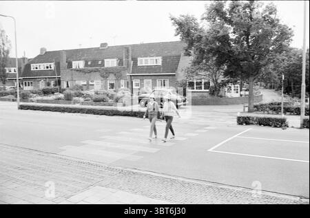 Zebra-Kreuzungen in Zandvoort Verkehr sicherer Verkehr, Whizgle News, Dutch Desk, Niederlande, 1950 - 2000 am 09-08-1963. Diese Themen sind in der Abbildung dargestellt. Die Szene fängt ein ruhiges urbanes Straßenbild ein, das von einem etwas bewölkten Himmel geprägt ist, der ein weiches, diffuses Licht über die Gegend wirft. Im Vordergrund gehen zwei Figuren zusammen und erscheinen in Gesprächen verwickelt. Ihre Kleidung erinnert an einen lässigen, modernen Stil, und sie bewegen sich über einen Fußgängerübergang. Auf beiden Seiten der Straße weisen Wohngebäude eine Mischung aus Backstein und Stuck auf. Die Strukturen, p Stockfoto