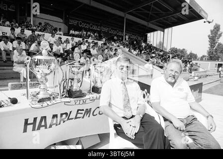 Haarlem Cup letzter Tag, Whizgle News, Dutch Desk, Niederlande, 1950 - 2000 am 07-08-1992. Die Abbildung zeigt diese Themen. Die Szene präsentiert eine lebhafte Outdoor-Umgebung, wahrscheinlich ein Sportereignis, mit einer Tribüne voller Zuschauer im Hintergrund. Die Ausdrucksformen des Publikums reichen von Begeisterung bis Vorfreude, was auf die Bedeutung des Ereignisses hinweist. Im Vordergrund sitzen zwei Individuen an einem Tisch, der mit mehreren Trophäen geschmückt ist, in Silber und Gold unter dem Licht. Die Trophäen sind unverwechselbar gestaltet und zeigen komplizierte Details und ein Gefühl für Prestige. Beide Männer sind gekleidet Stockfoto