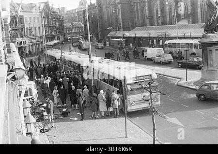Hektik Hartewens Festival Noord Zuid Hollandse Vervoersmaatschappij N.V. NZ, Whizgle News, Dutch Desk, Niederlande, 1950 - 2000 am 28-12-1964. Dies sind die Themen im Bild. Die Szene fängt eine geschäftige urbane Straße an einem kühlen Tag ein, gekennzeichnet durch eine Mischung aus Oldtimern und Menschenmassen. Im Vordergrund steht ein langer, knickgelenkter Bus mit weit geöffneten Türen, der die Passagiere zum Einsteigen einlädt. Menschen, die in einer Mischung aus Wintermänteln und Hüten gekleidet sind, versammeln sich auf dem Bürgersteig, einige steigen eifrig in den Bus, andere scheinen zu warten. Die Straße ist von VAR gesäumt Stockfoto