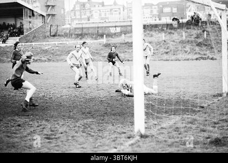 Zandvoort Meeuwen - EDO Football Zandvoortse Meeuwen Halfweg, Whizgle News, Dutch Desk, Niederlande, 1950 - 2000 am 18.04.1973. Diese Themen sind in der Abbildung dargestellt. Die Szene fängt einen intensiven Moment auf einem Fußballfeld in einer etwas düsteren Umgebung ein. Der Boden ist matschig, was auf jüngste Regenfälle hindeutet, mit einer unebenen, unebenen Textur unter den Füßen. Im Vordergrund breitet sich ein Torhüter in dunkler Uniform auf dem Boden aus und greift zum Ball, der in der Mitte der Bewegung nur wenige Zentimeter vom Torpfosten entfernt erfasst wird. Die Entschlossenheit des Torhüters steht im Gegensatz zur Herausforderung Stockfoto