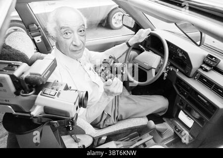 Erfinder Gatsonides Overveen Inventions Overveen, Whizgle News, Dutch Desk, Niederlande, 1950 - 2000 am 21-07-1986. Diese Themen sind in der Abbildung dargestellt. Die Szene zeigt einen älteren Mann, der in einem Auto sitzt, in einem Moment der Konzentration und Nostalgie verwickelt ist. Der Innenraum des Fahrzeugs zeichnet sich durch eine Mischung aus gedämpften Grau- und sanften Beigetönen aus, die eine eher Vintage-Atmosphäre vermitteln. Die Kleidung des Mannes besteht aus einem hellen Hemd, das sich von den dunkleren Elementen des Armaturenbretts des Autos abhebt. Er hält eine alte Kamera in der rechten Hand, während ein größerer Camcorder auf dem Armaturenbrett in der Nähe liegt. Beides Stockfoto