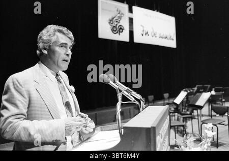 Congress Print and Paper Concert Hall Congress, Whizgle News, Dutch Desk, Niederlande, 1950 - 2000 am 26-05-1988. Dies sind die Elemente im Bild. In einem schwach beleuchteten Auditorium steht ein Mann an einem hölzernen Podium und spricht in mehrere Mikrofone, die in der Nähe positioniert sind. Er trägt einen hellen Anzug mit einer blumigen Krawatte, die der Szene einen Hauch von Lebendigkeit verleiht. Sein Haar ist zurückgeschliffen, und er hat einen konzentrierten Ausdruck, der ein Gefühl von Autorität und Engagement für das Publikum vermittelt. Hinter ihm hängen Banner von der Decke, deren Design auf künstlerische Elemente oder Themen im Zusammenhang mit der Veranstaltung hinweist. Stockfoto