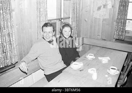 Könnte. Familie im Urlaub (Gstaad) Kon. Besuche und Empfänge Personen Beerdigung, Whizgle News, Dutch Desk, Niederlande, 1950 - 2000 am 22-01-1966. Dies sind die Elemente im Bild. Die Szene zeigt einen gemütlichen Essbereich mit warmen Holzwänden und -Möbeln, die zu einer einladenden, rustikalen Atmosphäre beitragen. Natürliches Licht filtert durch große Fenster und beleuchtet den Bereich hell. Im Vordergrund sitzt ein Paar bequem an einem langen Holztisch. Der Mann, der lässig in einem leichten Pullover gekleidet ist, zeigt einen fröhlichen Ausdruck, der Wärme und Leichtigkeit hervorbringt. Er neigt sich leicht zu seinem Co Stockfoto